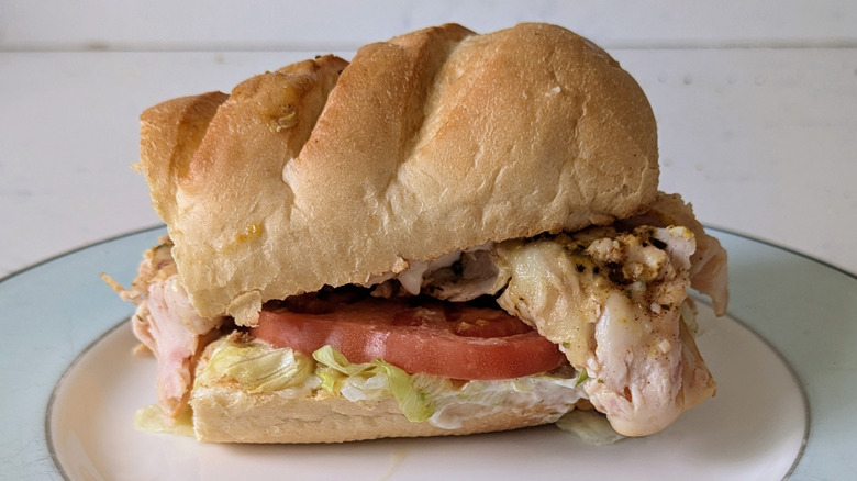 A sub hoagie roll is sliced in half and topped with classic toppings including tomato, mayo, lettuce, and turkey with some kind of seasoning evident on the turkey and cheese.