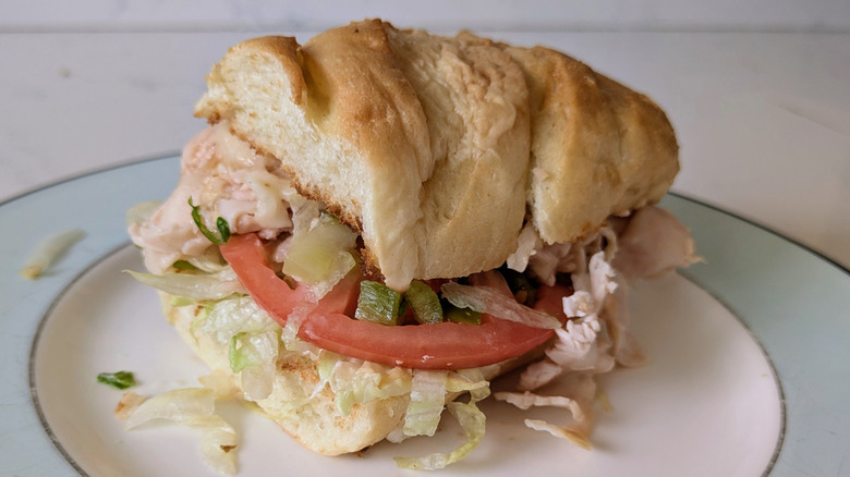 A toasted white sub hoagie roll on a blue and white plate features a sliced tomato, chopped pickles, turkey, provolone cheese