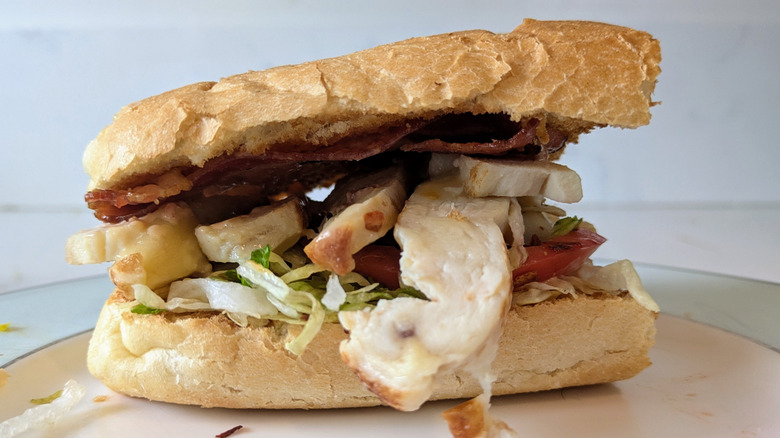 A toasted white sub hoagie roll is sliced in half and topped with shredded green lettuce, grilled chicken breast, sliced tomato, strips of bacon, on a white and blue plate