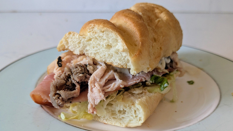 A toasted white sub hoagie roll is sliced in half and topped with ham, roast beef, turkey, shredded green lettuce on a white and blue plate