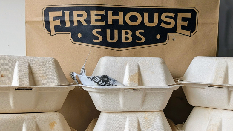 A brown grocery bag with a black Firehouse Subs logo stands behind six paper-carton to-go sub sandwich boxes