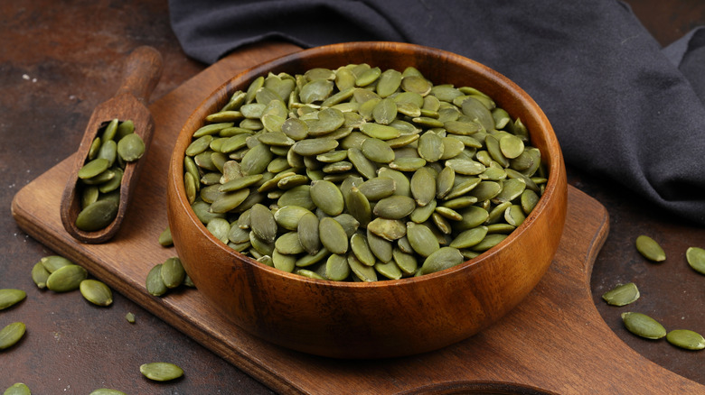Pumpkin seeds in a wooden bowl on a cutting board.