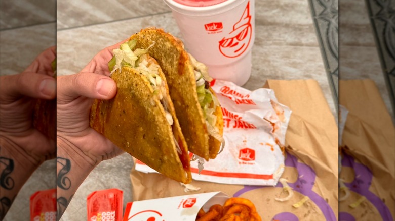 A hand holding two Jack in the Box tacos over some paper wrappers with a drink
