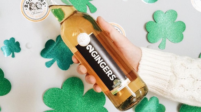 A person holding a bottle of 2 Gingers whiskey with cut out clovers in the background