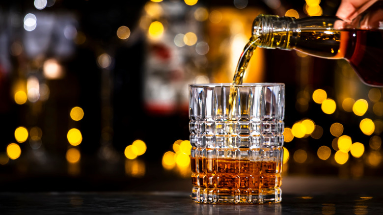 A bottle of whiskey being poured into a glass with blurred lights in the background