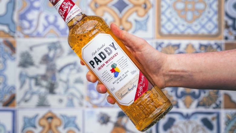 A person holding a bottle of Paddy Irish whiskey to the camera in front of a colorful blurred background