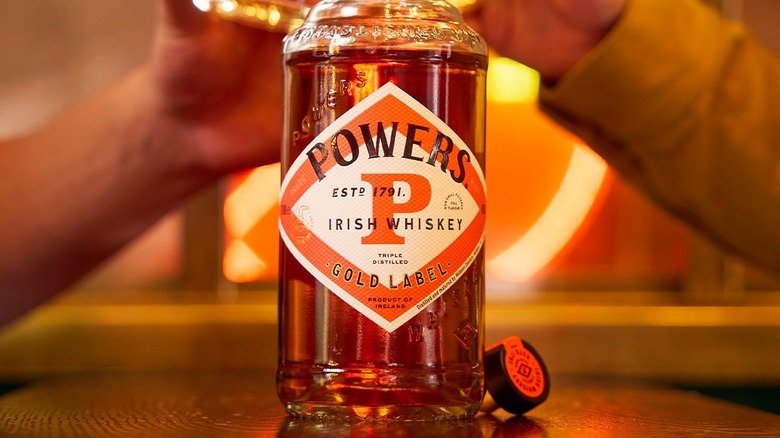 A bottle of Powers whisky some in the foreground with people clinking glasses in the background