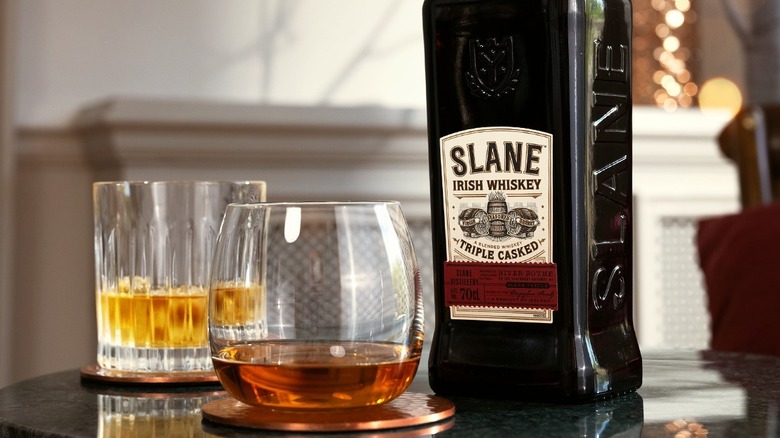 A bottle of Slane Irish Whiskey on a table next to two glasses, one neat whiskey and the other a cocktail