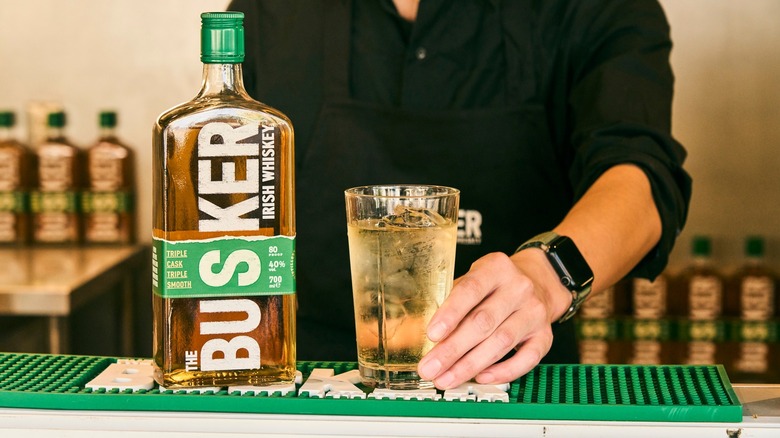 A bottle of The Busker on the side of a bar next to a cocktail with a bartender in the background