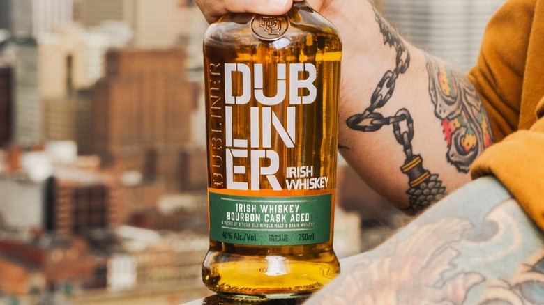 A tattooed person holding a bottle of The Dubliner to the camera with a blurred city background
