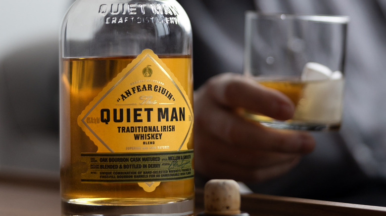 A close up of a bottle of The Quiet Man with a person holding a filled glass in a blurry background
