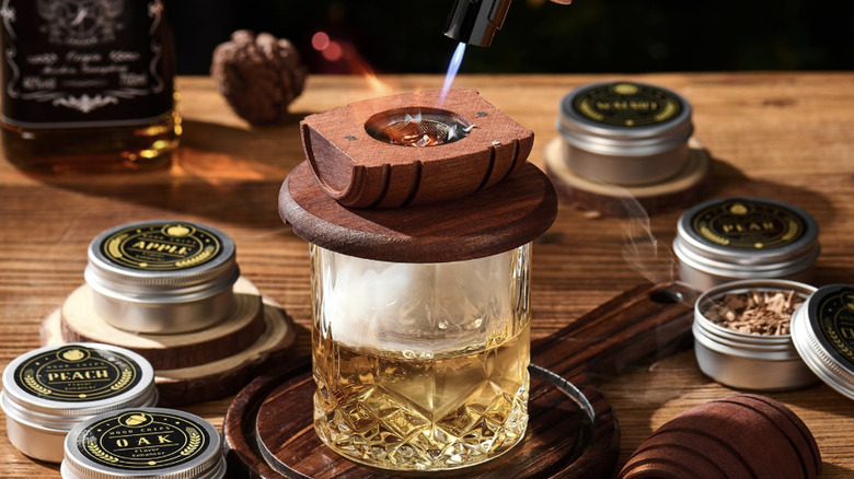Using whiskey smoker kit to infuse glass of bourbon next to metal containers holding wood chips