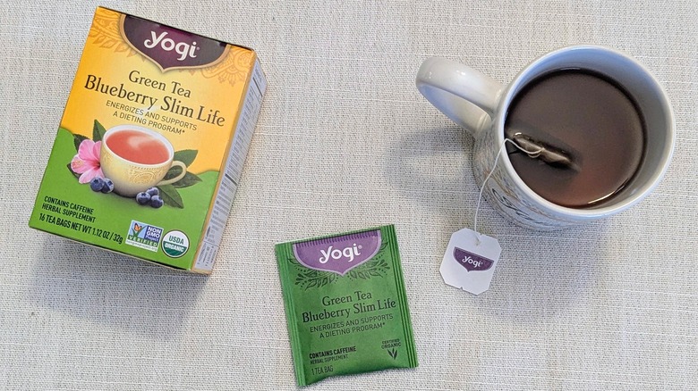 A box of Green Tea Blueberry Slim Life tea, a tea bag, and a mug on a white tablecloth