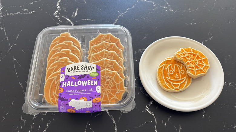 A container of Bake Shop Halloween Sugar Cookies next to two sugar cookies, one bitten, on a white plate