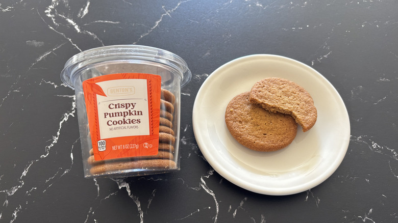 A container of Benton's Crispy Pumpkin Cookies next to two cookies, one bitten, on a white plate
