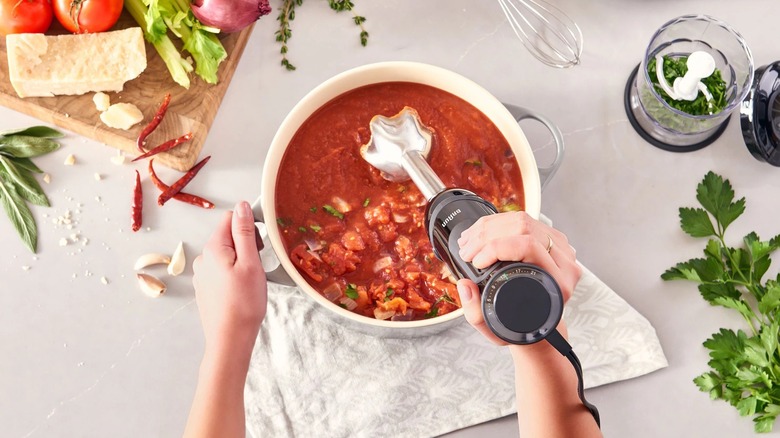 A person using a hand blender to mix pasta sauce in a large pot on a counter surrounded by ingredients