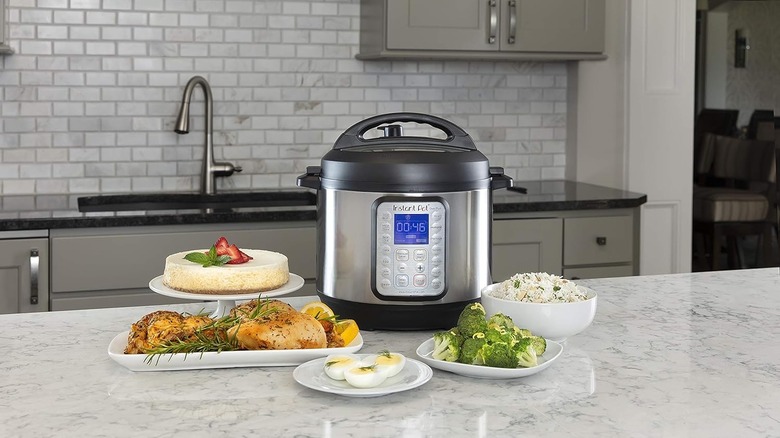 An instant Pot pressure cooker with a blue digital display sitting on a counter next to a cheesecake, vegetables, cooked meat, boiled eggs and a bowl of rice