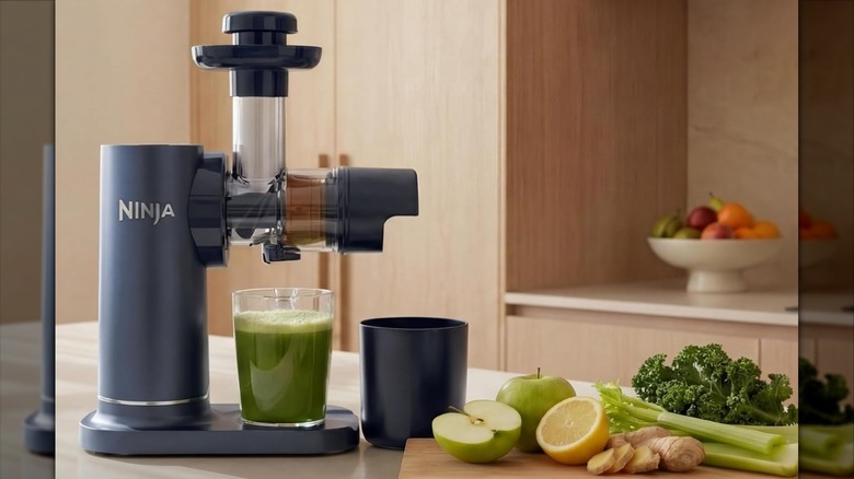 A dark gray Ninja juicer with a glass of green juice under it and some green veggies and fruits on the side