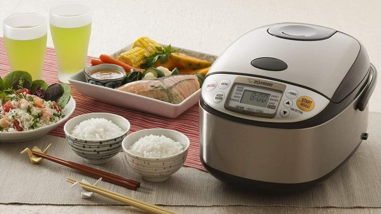 A rice cooker on a table next to several bowls of rice, chopsticks, plated veggies, and drinks