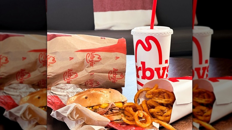 An Arby's Beef and Cheddar sitting on paper next to curly fries and an Arby's cup