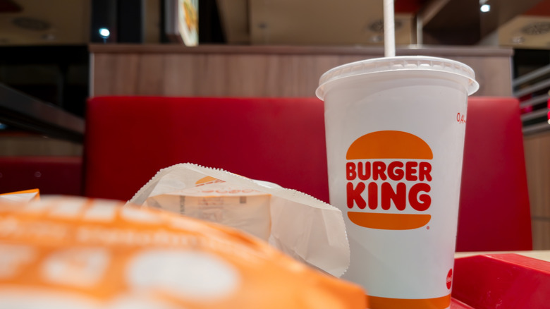 A Burger King drink cup to the right side of a tray with a wrapped Burger King burger out of focus in the foreground