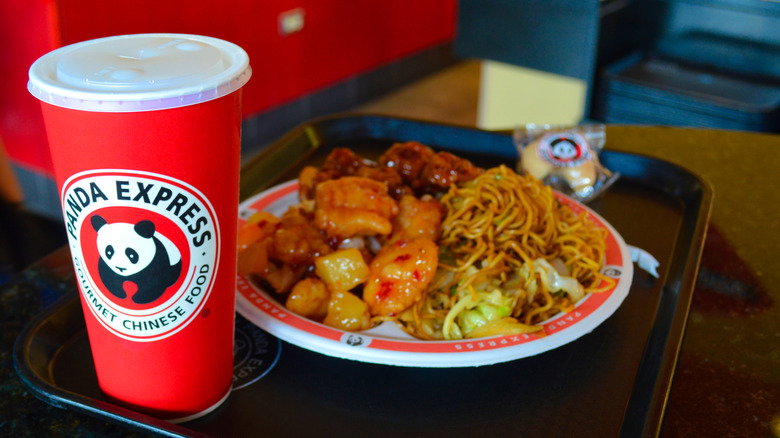 A plate of orange chicken and noodles next to a Panda Express drink cup