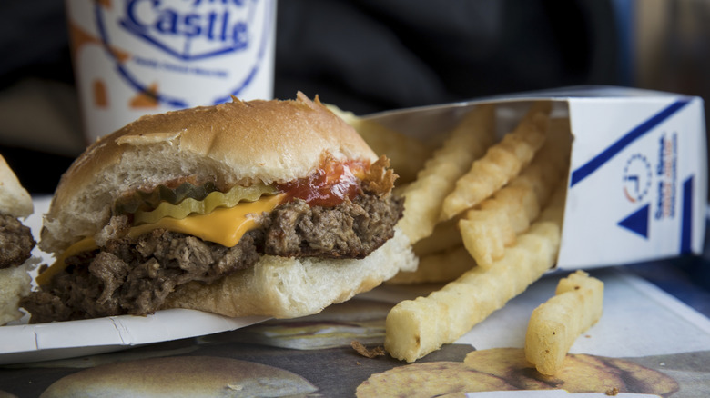 Close up of a White Castle cheeseburger with visible pickles and ketchup next to some crinkle cut fries and a drink in the background
