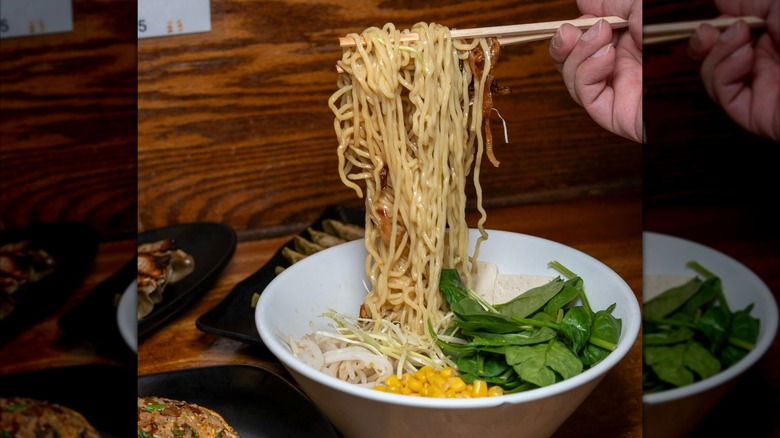 Hand holding ramen noodles on chopsticks over bowl of corn and greens