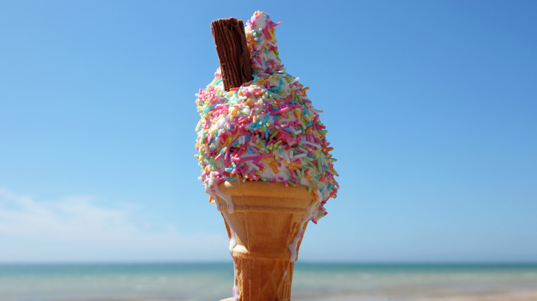 Ice cream covered in sprinkles in a sugar cone with a piece of chocolate sticking out from the left side.