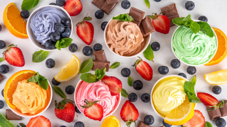 Cups of frozen yogurt with orange, blueberry, strawberry, chocolate, lemon, and mint flavors surrounded by scattered ingredients of each flavor.