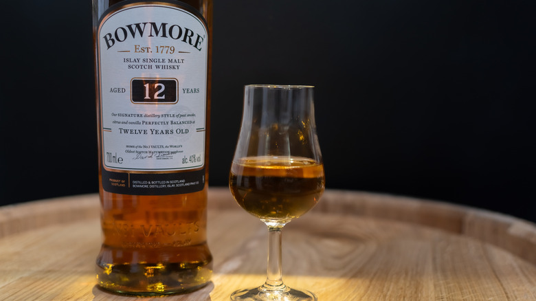 A bottle of Bowmore 12-year on top of a barrel next to a filled glencairn glass