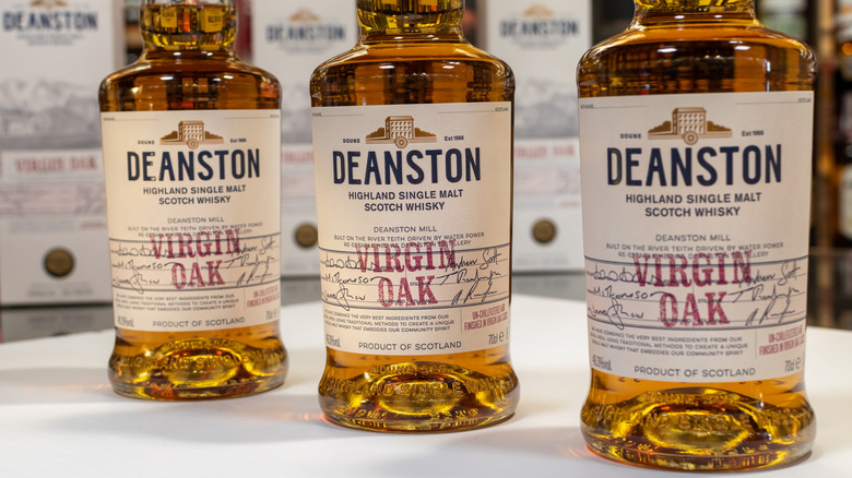 Three bottles of Deanston Virgin Oak single malt whisky posed side-by-side on a table with blurred boxes in the background