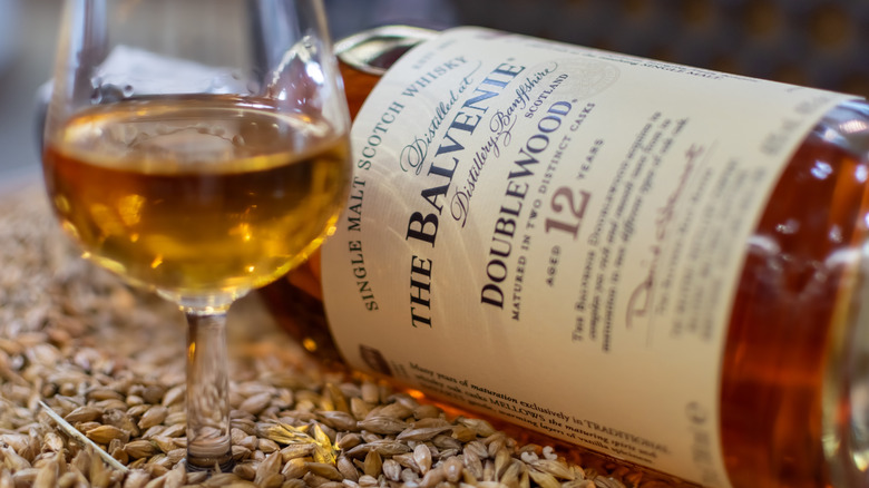 A bottle of The Balvenie DoubleWood single malt scotch lying on its side on grain next to a filled glass