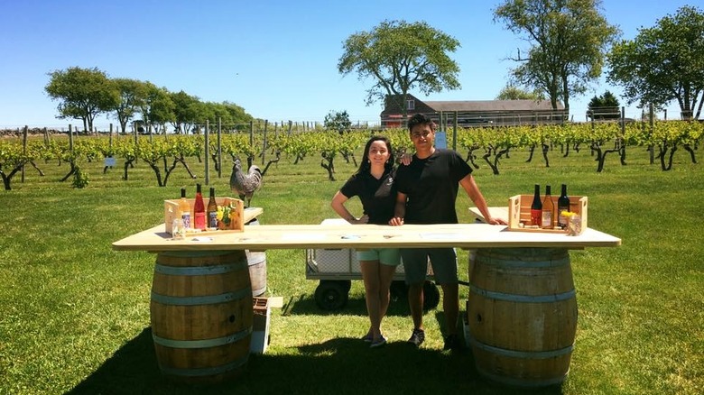 A man and a woman operating a wine tasting table, with a winery and vineyard behind them
