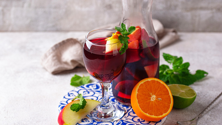 A jug and glass of sangria with orange, apple and lime pieces around it