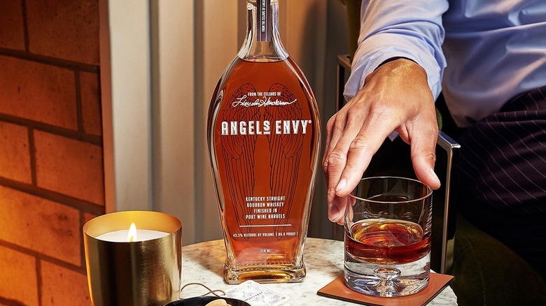 A bottle of Angel's Envy place don a table with a person reaching for a filled glass next to it