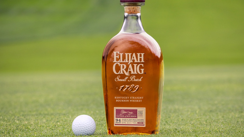 A bottle of Elijah Craig bourbon posed on a golf green with a golf ball next to it