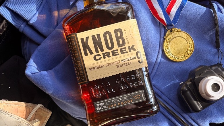 A bottle of Knob Creek laid on an item of clothing with a toy camera and medal next to it
