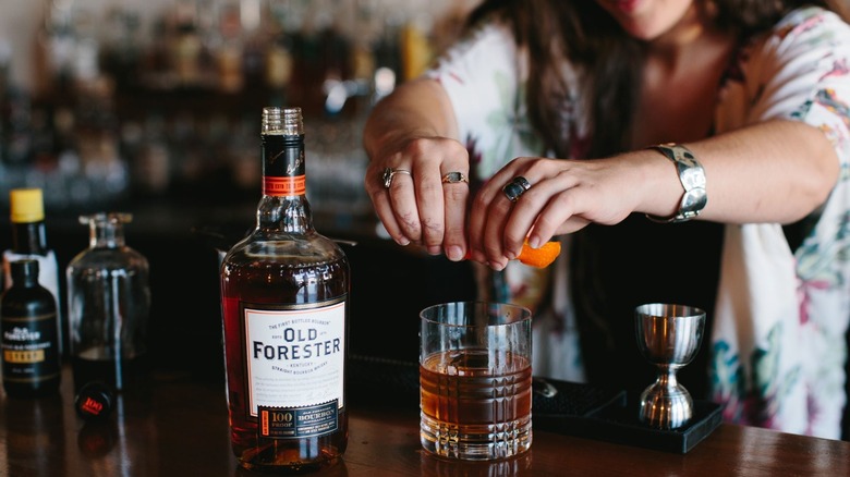 A bottle of Old Forester 100 Proof on a bar with a bartender making a cocktail next to it