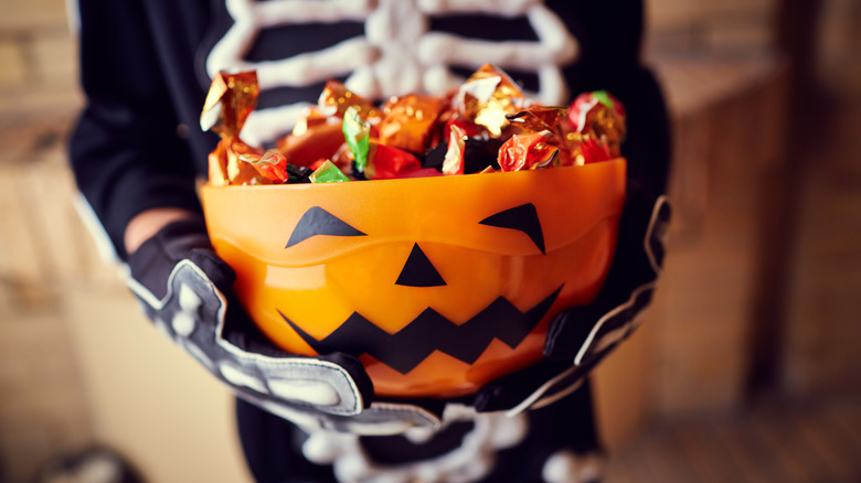 Person dressed as skeleton holding Jack-o-Lantern candy bucket filled with candy for Halloween