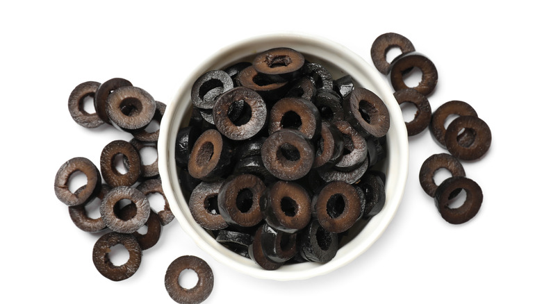 Sliced black olives in a white container with some surrounding it, with a white background.