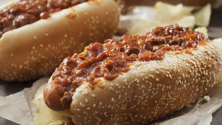 Two hot dogs topped with chili on parchment paper with potato chips.