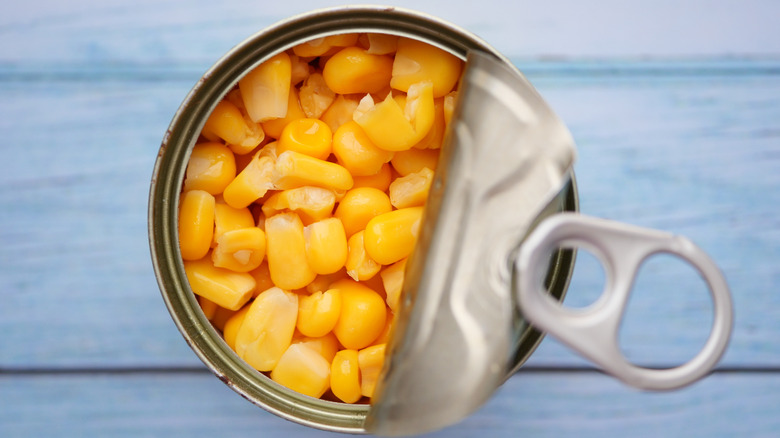 A partially opened can of corn kernels on a light bluish white background.