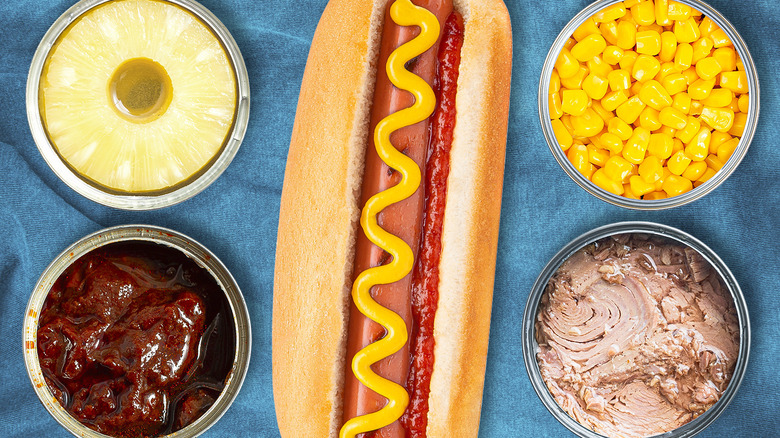 A hot dog dressed with ketchup and mustard, surrounded by cans of sliced pineapple, chipotle peppers in adobo sauce, corn kernels, and tuna on a blue tablecloth.