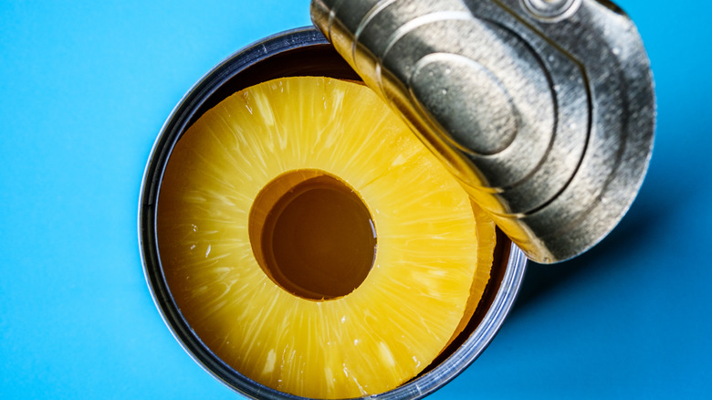 Pineapple rings in a can with the lid partially peeled off, with a blue background.