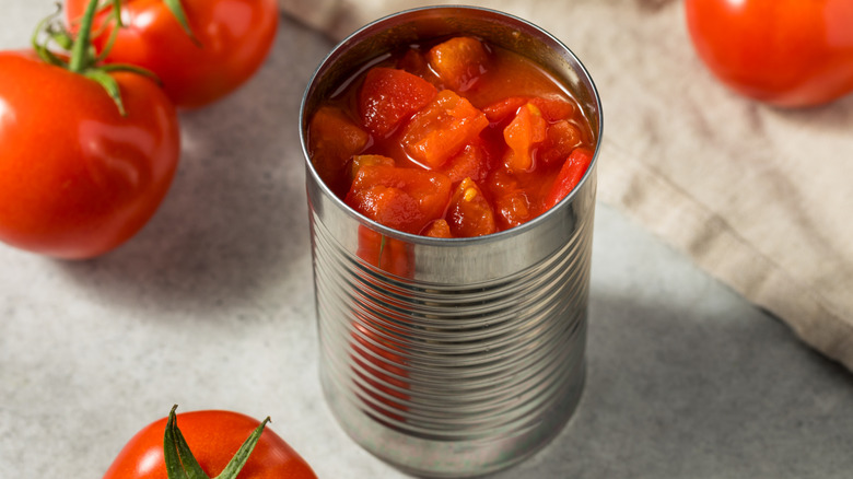 Diced tomatoes in a tin can, on a table surrounded by fresh tomatoes.