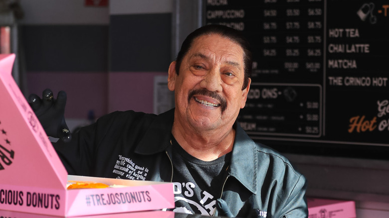 Danny Trejo smiling while holding a pink box of Trejo's Donuts