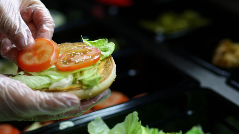 Gloved hands placing a tomato on a burger bun with lettuce