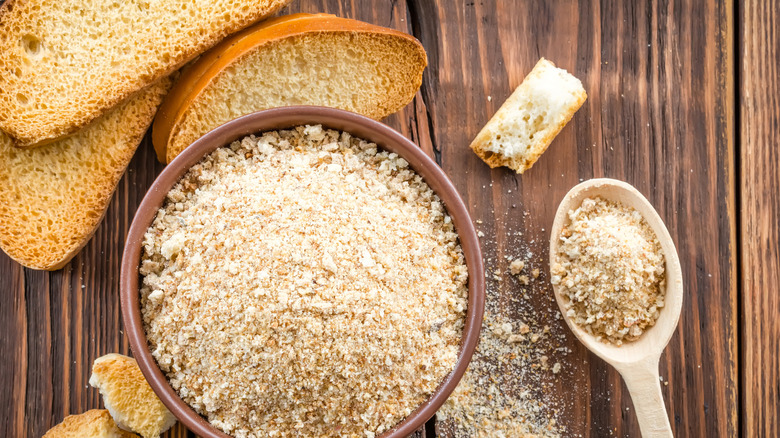 A bowl full of bread crumbs on a wooden table next to some slices of toasted bread and a spoonful of crumbs