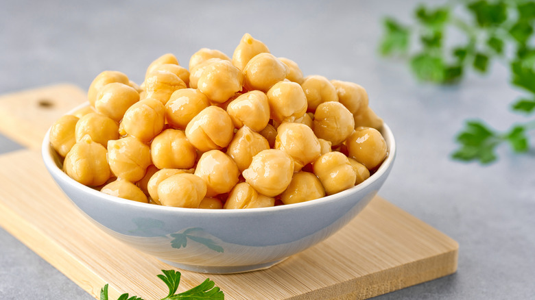 A white bowl of chickpeas sitting on a wooden cutting board with some parsley in the foreground and background